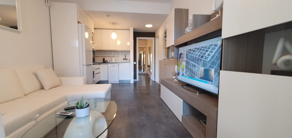 Sala de estar moderna con sofá, mesa de cristal y cocina abierta al fondo.